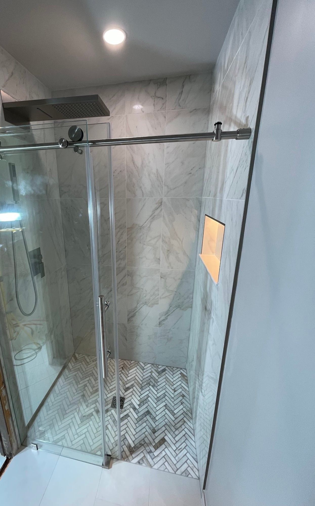 Frameless glass shower in marble-look tile with a herringbone floor and lit niche